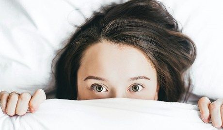 Frau blickt mit großen Augen unter der Bettdecke hervor